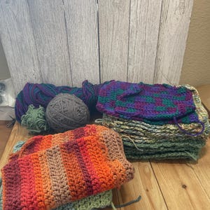 May include: A collection of colorful crochet projects and yarn balls. The projects include a stack of green and purple squares, and a vibrant orange and red striped piece. Several balls of yarn in shades of purple, green, and gray are also present.