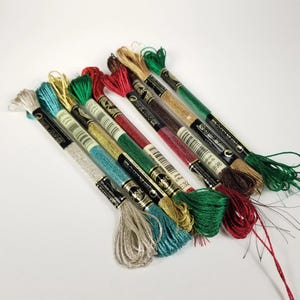 May include: A collection of embroidery floss in various colors, including green, red, gold, and teal. The spools are arranged diagonally against a white background, showcasing the different shades and textures of the thread. Ideal for embroidery and cross-stitch projects.