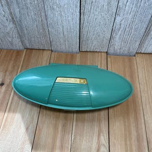 May include: An oval-shaped, teal-colored plastic storage case with a hinged lid. The center of the lid features a gold-colored rectangular plate with the word SINGER embossed in black lettering, set against a ribbed, horizontal textured panel.