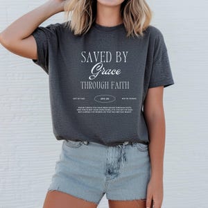 May include: A gray t-shirt with the text "SAVED BY Grace THROUGH FAITH" in white. The text is surrounded by a white border and includes the scripture reference "EPH. 2:8".