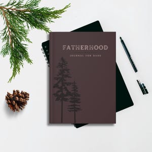 May include: A maroon journal with the word "FATHERHOOD" and "JOURNAL FOR DADS" printed on the cover. Two pine tree silhouettes are below the text. A black pen, pine cone, and evergreen branch are also in the image.