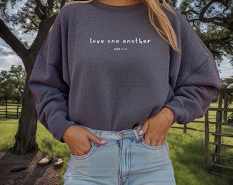 以下が含まれることがあります： 白い文字で「love one another」と「JOHN 13:34」と書かれた濃いグレーのトレーナーです。