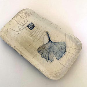 May include: A rectangular ceramic tray with a cream-colored crackle glaze finish. The surface features a blue ginkgo leaf illustration, delicate line drawings of foliage, and a small square blue stamp with Asian characters in the upper left corner.