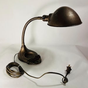 Puede incluir: Una lámpara de escritorio de metal en tono bronce con una pantalla en forma de cúpula, un brazo flexible de cuello de cisne y una base decorativa con una hendidura circular y un soporte para bolígrafos. Incluye un cable eléctrico largo.