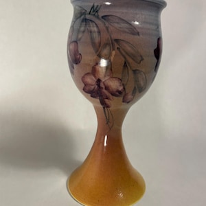 May include: A ceramic goblet featuring a hand-painted floral design with dark purple and maroon blossoms on a gradient background that transitions from a soft lavender at the rim to a warm, golden-orange hue at the flared circular base.