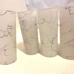May include: Four frosted glass tumblers with black abstract line art. The glasses are cylindrical and have a slightly textured surface. The black lines create a free-flowing, artistic design on the white background.