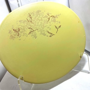 Könnte beinhalten: Ein runder, hellgelber dekorativer Teller mit einem goldenen Blattdesign. Der Teller wird von einem transparenten Acrylständer getragen. Das Blattdesign ist in der Mitte gruppiert und um den Teller verstreut.