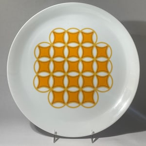 Könnte beinhalten: Ein weißer Keramikteller mit einem geometrischen Muster aus überlappenden Kreisen und Quadraten in Orange- und Gelbtönen.