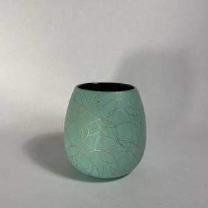 May include: A rounded, bulbous cup with a matte mint green exterior featuring a delicate, thin gold-colored line pattern of intersecting loops and swirls. The interior of the cup is a solid, contrasting black, creating a distinct visual edge.
