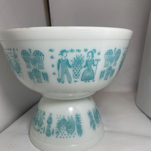 May include: White pedestal bowl with a turquoise design featuring figures, baskets, and plants. The bowl has a wide top and a smaller, rounded base. The turquoise design wraps around the bowl, creating a vintage aesthetic.