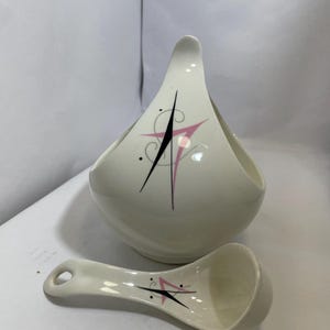 May include: Cream-colored ceramic serving bowl with a handle and matching spoon. The bowl and spoon feature a mid-century modern design with pink and black geometric accents. The bowl is approximately 7 inches wide.