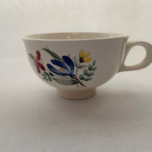 May include: Cream-colored teacup with a floral design featuring blue, yellow, and pink flowers with green leaves. The teacup has a rounded bowl shape, a handle, and a small base.