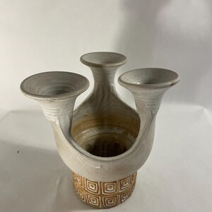 May include: A ceramic vase with three distinct flared openings extending from a central body. The upper portion is finished in a matte off-white glaze, while the lower base features a repeating geometric square pattern in shades of brown and white.