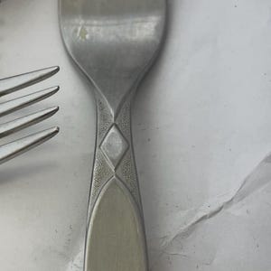 Puede incluir: Primer plano de un tenedor plateado con cuatro púas. El mango tiene un diseño de diamantes decorativo. La cubertería es probablemente parte de un juego de comedor.