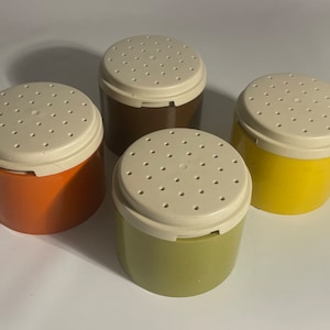 May include: Four colorful spice shakers. Each shaker has a cylindrical base in orange, brown, yellow, and green. The lids are off-white with small holes. The shakers are likely made of plastic and are designed for dispensing spices or seasonings.