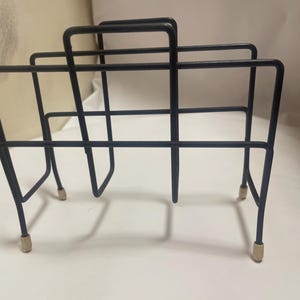 May include: A black metal magazine rack with a rectangular design. The rack has a three-sectioned design with a handle on top. The legs have small, gold-colored feet. The rack is designed to hold magazines or other reading materials.