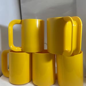 May include: A stack of five bright yellow mugs. Each mug has a rounded, rectangular handle. The mugs are made of a solid, glossy material and are evenly lit, showcasing their simple, modern design. The mugs are stacked on top of each other.