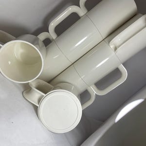 May include: A collection of off-white, cylindrical mugs with large, rectangular handles. The image shows several mugs stacked together, along with lids and a single mug. The mugs appear to be made of a durable plastic material.