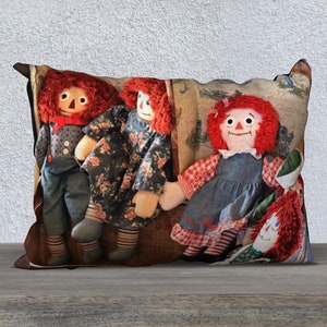 May include: A pillow featuring a colourful fabric design with four red-haired rag dolls. The dolls are wearing different outfits, including a blue dress, a floral dress, and a blue shirt and trousers. The pillow is ideal for adding a touch of whimsy to any room.