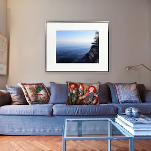 May include: A framed photograph of a lake with a cliff in the background. The lake is a light blue colour and the cliff is a dark grey colour. The photograph is hanging on a wall in a living room with a blue sofa and cushions.