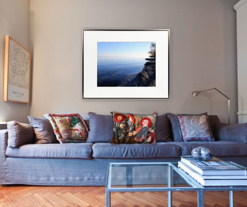 May include: A framed photograph of a lake with a cliff in the background. The lake is a light blue colour and the cliff is a dark grey colour. The photograph is hanging on a wall in a living room with a blue sofa and cushions.