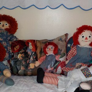 May include: A group of five red-haired rag dolls sitting on a white and blue quilted surface. The dolls are wearing various blue and white patterned dresses and have red and white striped stockings.