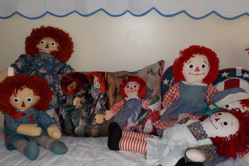May include: A group of five red-haired rag dolls sitting on a white and blue quilted surface. The dolls are wearing various blue and white patterned dresses and have red and white striped stockings.