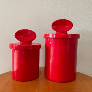 Puede incluir: Dos botes de cerámica rojos con tapas. Las tapas tienen una forma ovalada redondeada y están unidas a los botes. Los botes son de diferentes tamaños.