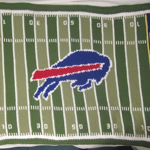 May include: Crocheted football field blanket with a blue and red Buffalo Bills logo in the center. The blanket has the words "Bills" on each side of the logo.