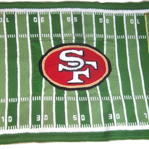 May include: A green and white knitted blanket with a football field design. The center of the blanket features a red and gold San Francisco 49ers logo. The words "49ers" are on the sides of the blanket.