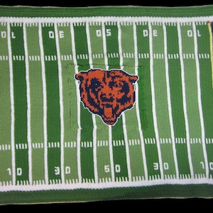 May include: A rectangular crochet blanket featuring a green football field design with white yard lines and numbers. The text CHICAGO is on the left and BEARS is on the right in orange letters on navy blue panels. A bear head logo is in the center.