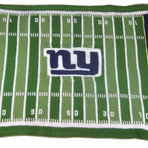 May include: A handmade, crocheted blanket in the style of an American football field. The blanket features green and white stripes, with the "NY GIANTS" logo and team name in blue and white. The field markings and end zones are clearly defined.