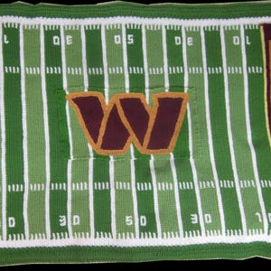 Puede incluir: Una manta de ganchillo con un diseño de campo de fútbol americano. La manta es verde con líneas blancas y el nombre del equipo "COMMANDERS" en letras doradas sobre un borde burdeos. Una gran "W" burdeos está centrada.