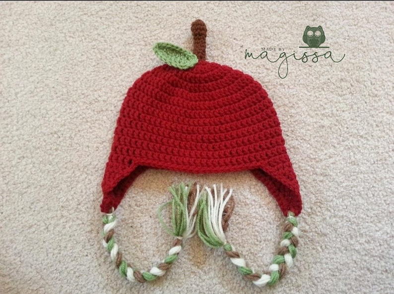 May include: Red crocheted apple hat with green leaf and brown, white, and green braided ties. "Made by magissa" is written in the top right corner of the image.