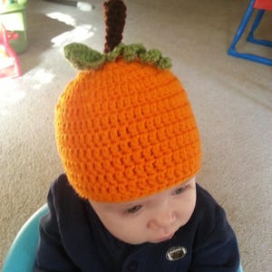 May include: A baby wearing a crocheted orange pumpkin hat with green leaves and a brown stem. The baby is wearing a navy blue shirt and is sitting in a blue high chair.