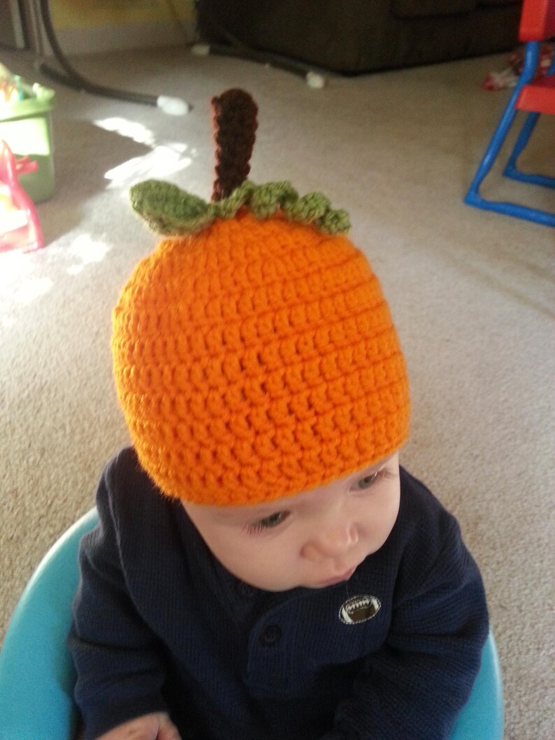May include: A baby wearing a crocheted orange pumpkin hat with green leaves and a brown stem. The baby is wearing a navy blue shirt and is sitting in a blue high chair.