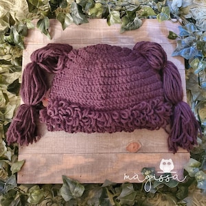 May include: A plum-colored crochet hat with attached yarn braids. The hat features a textured stitch and a ruffled edge. The braids are thick and hang down on either side. The hat is displayed on a wooden surface with greenery.