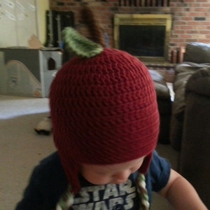 May include: A red crocheted hat shaped like an apple with a green leaf on top. The hat has ear flaps and is worn by a child wearing a blue shirt with the words "Star Wars" printed on it.