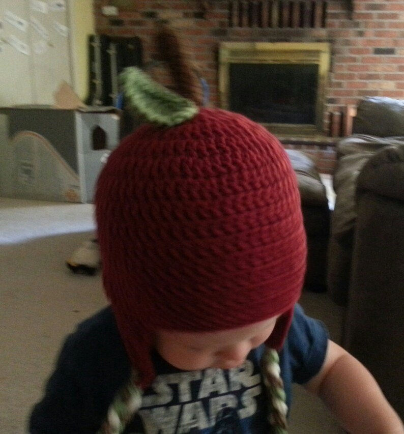 May include: A red crocheted hat shaped like an apple with a green leaf on top. The hat has ear flaps and is worn by a child wearing a blue shirt with the words "Star Wars" printed on it.