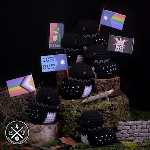 May include: Handmade crochet loon figurines with black bodies, white bellies, and red eyes. Each loon holds a miniature flag with various designs, including a rainbow flag and the words "NO KINGS". The figurines are displayed on a mossy surface.