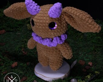 May include: A brown crocheted plush creature with large, floppy ears, two small purple horns, and a purple ruff around its neck. The creature has large, dark purple eyes and stands on a circular white base. A logo with the text 4 H T is visible.