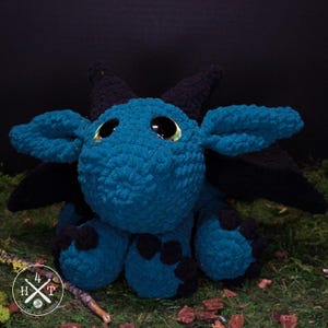 May include: A teal crocheted dragon plushie with black horns, wings, and feet. The dragon has large, round, yellow-rimmed eyes. The plushie is sitting on a bed of green moss.