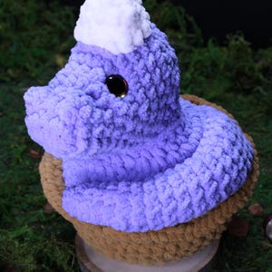 May include: A handmade, plush, purple mushroom-shaped item with a white top, brown base, and a large black eye. The mushroom is sitting on a wooden disc, surrounded by green moss.