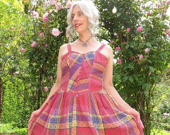 May include: A woman wearing a pink, blue, and yellow plaid dress with a pink trim. The dress has a fitted bodice and a full skirt. She is standing in a garden with pink roses.