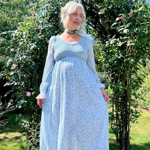 May include: A long, light blue dress with a floral design. The dress has long sleeves with lace cuffs, a smocked bodice, and a tiered skirt. The dress is worn in a garden setting, with green foliage in the background.