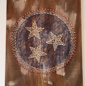 May include: String art of the state of Tennessee flag on a wooden board. The flag is a white circle with three red stars on a blue background. The circle is surrounded by a red and white border.