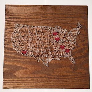 May include: A string art map of the United States with red hearts marking locations on a brown wooden background.