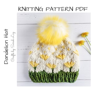 May include: A white knitted hat with a yellow dandelion pattern and a green brim. The hat has a yellow and white pom pom on top. The hat is on a white wooden surface.