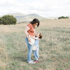 May include: A woman and a young girl are standing in a field, smiling and hugging. The woman is wearing a red top and blue jeans, and the girl is wearing a blue shirt and jeans. They are both wearing white crocheted bags.