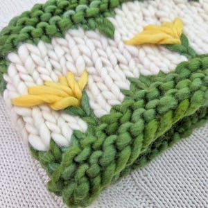 Puede incluir: Un gorro de punto grueso en crema, verde y amarillo. El gorro presenta un patr&oacute;n repetido de flores amarillas con tallos y hojas verdes sobre un fondo crema. El borde y los contornos de las flores son verdes.